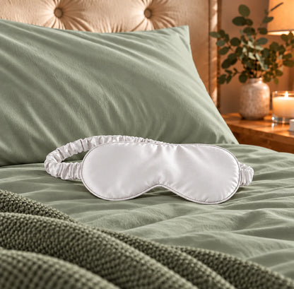 White bamboo lyocell sleep eye mask with a ruched strap on sage green bedding in a cosy bedroom with a tufted headboard and bedside candle.