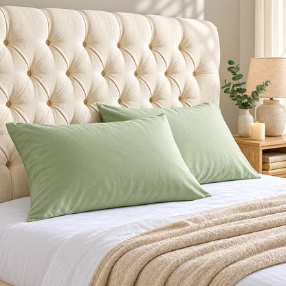 Two sage green bamboo lyocell pillowcases styled on a white bed with an ivory tufted headboard, beige textured throw and neutral bedside decor.